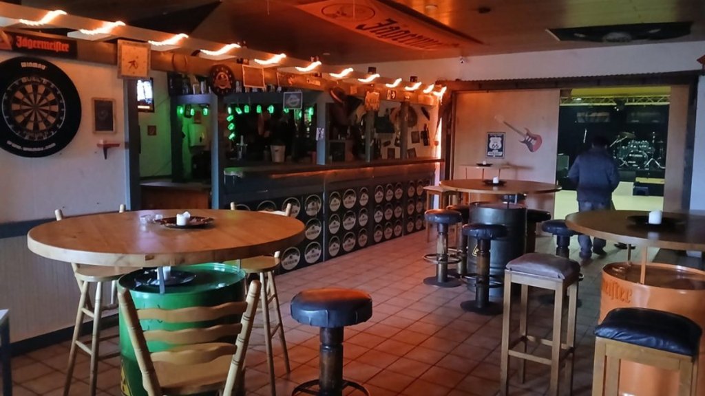 Innenraum einer Bar mit mehreren runden Holztischen, Barhockern und einer Dartscheibe an der Wand.