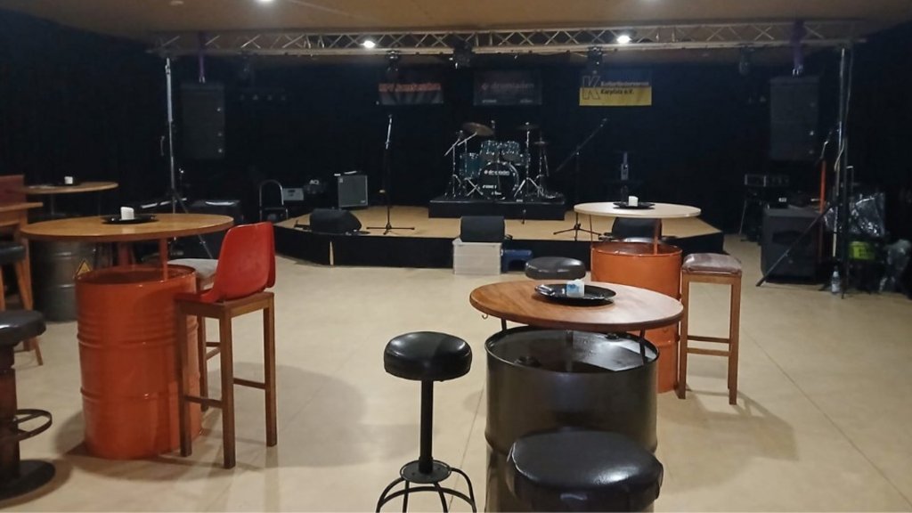 Leerer Veranstaltungsraum mit Bühne, Schlagzeug, Lautsprechern und mehreren Tischen mit Hockern und Stühlen.