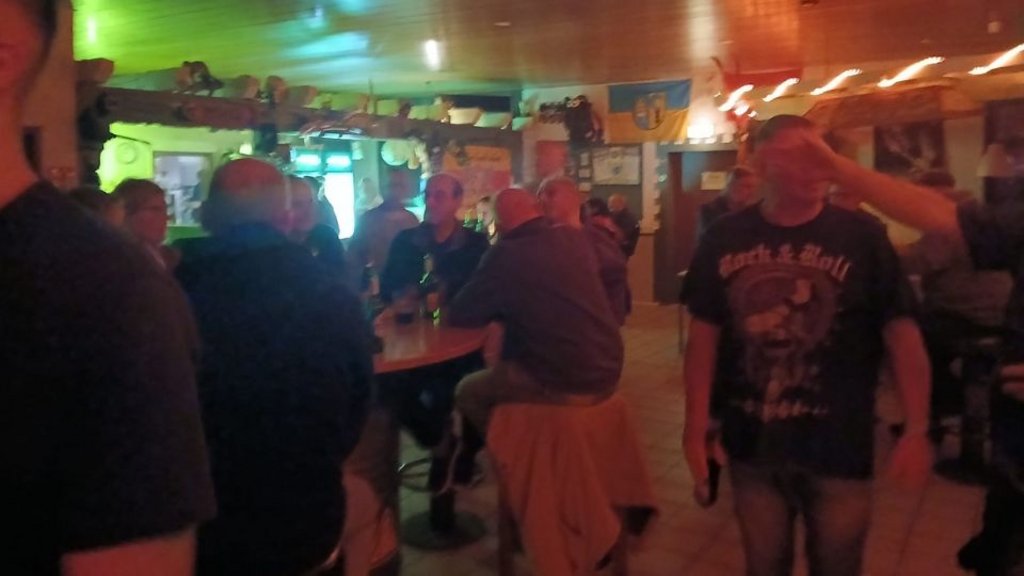 Mehrere Personen sitzen und stehen in einem rustikalen Gastraum mit Holzwänden und Decke, einige tragen dunkle Kleidung.