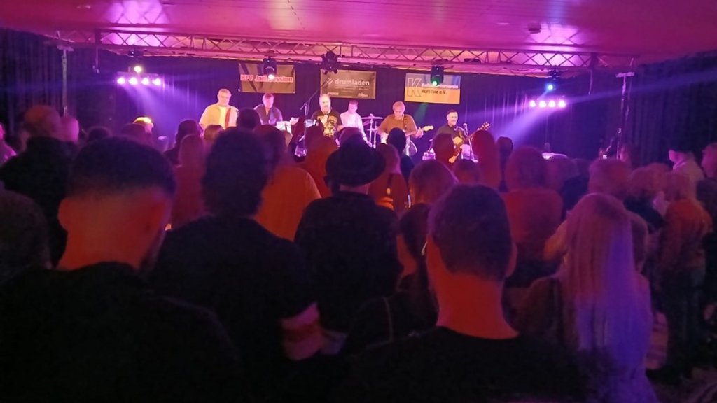 Band mit sechs Musikern spielt auf Bühne vor Publikum in dunklem Raum mit lila Bühnenbeleuchtung.